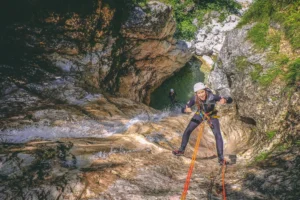 Woman canyoning down a rocky waterfall using ropes, wearing a helmet and wetsuit, giving a thumbs-up with a big smile.