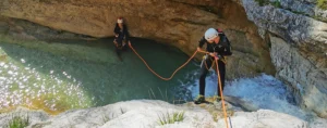 Two canyoners in wetsuits and helmets navigating a narrow canyon; one person rappelling down beside a waterfall while the other stands in a turquoise pool holding the safety rope