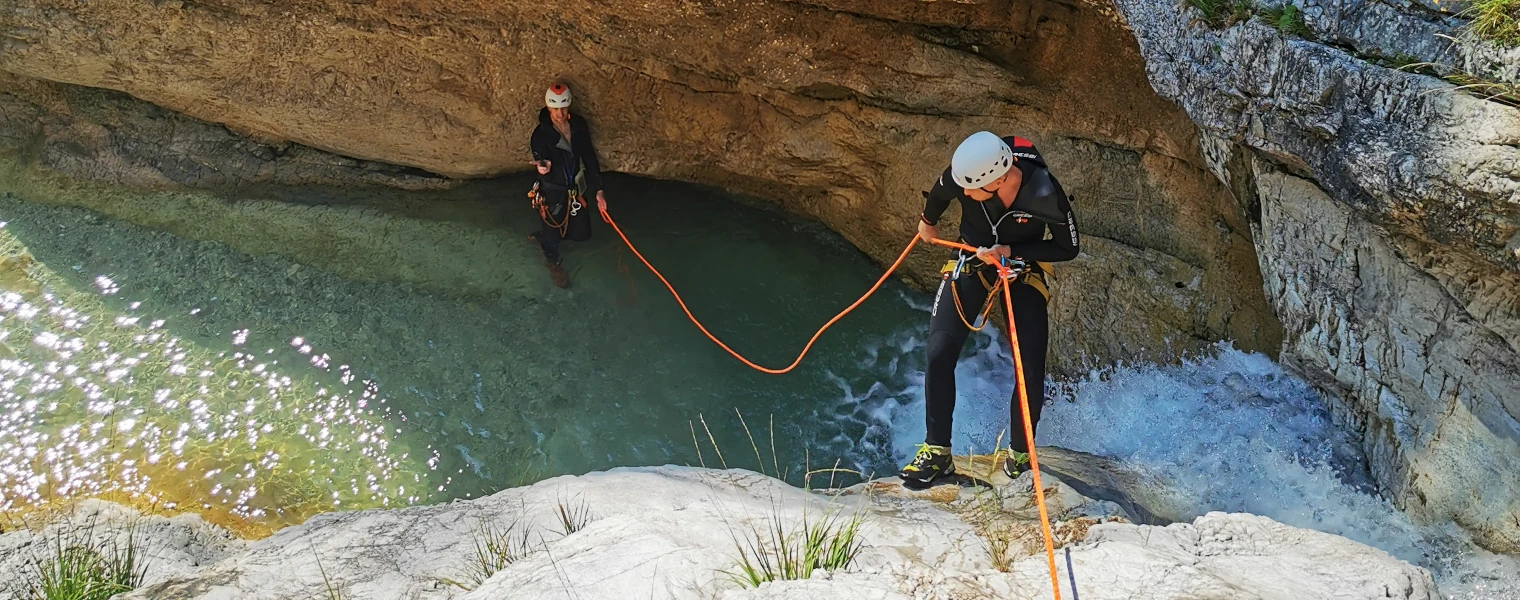 Two canyoners in wetsuits and helmets navigating a narrow canyon; one person rappelling down beside a waterfall while the other stands in a turquoise pool holding the safety rope