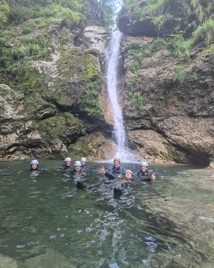 Group of canyoneers wearing helmets and wetsuits standing in a pool beneath a tall waterfall in Sušec Canyon.